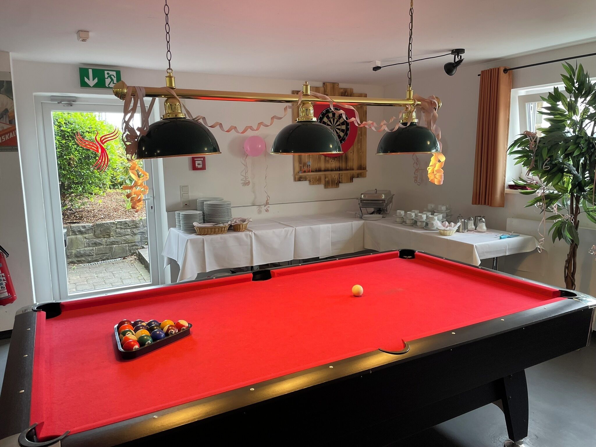 A pool table with balls, a dartboard, and a table with cups and condiments.