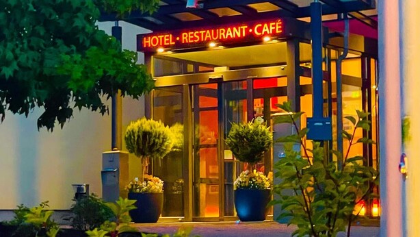 A hotel with a glass facade, a sign reading 'HOTEL RESTAURANT CAFÉ,' and potted plants in front. - PHÖNIX Hotel (Bergneustadt)