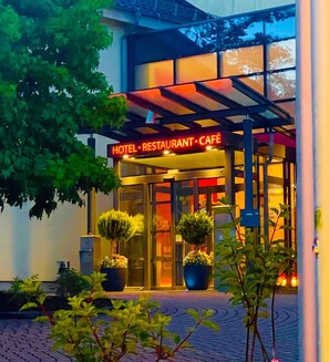 A hotel with a glass facade, a sign reading 'HOTEL RESTAURANT CAFÉ,' and potted plants in front. - PHÖNIX Hotel (Bergneustadt)