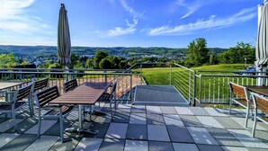 An outdoor patio with tables and benches, a metal railing, and a view of a town and hills in the distance. - PHÖNIX Hotel (Bergneustadt)
