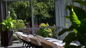 A sunlit room with a view of lush greenery through large windows, a potted plant, and a row of sun loungers with towels. - PHÖNIX Hotel (Bergneustadt)
