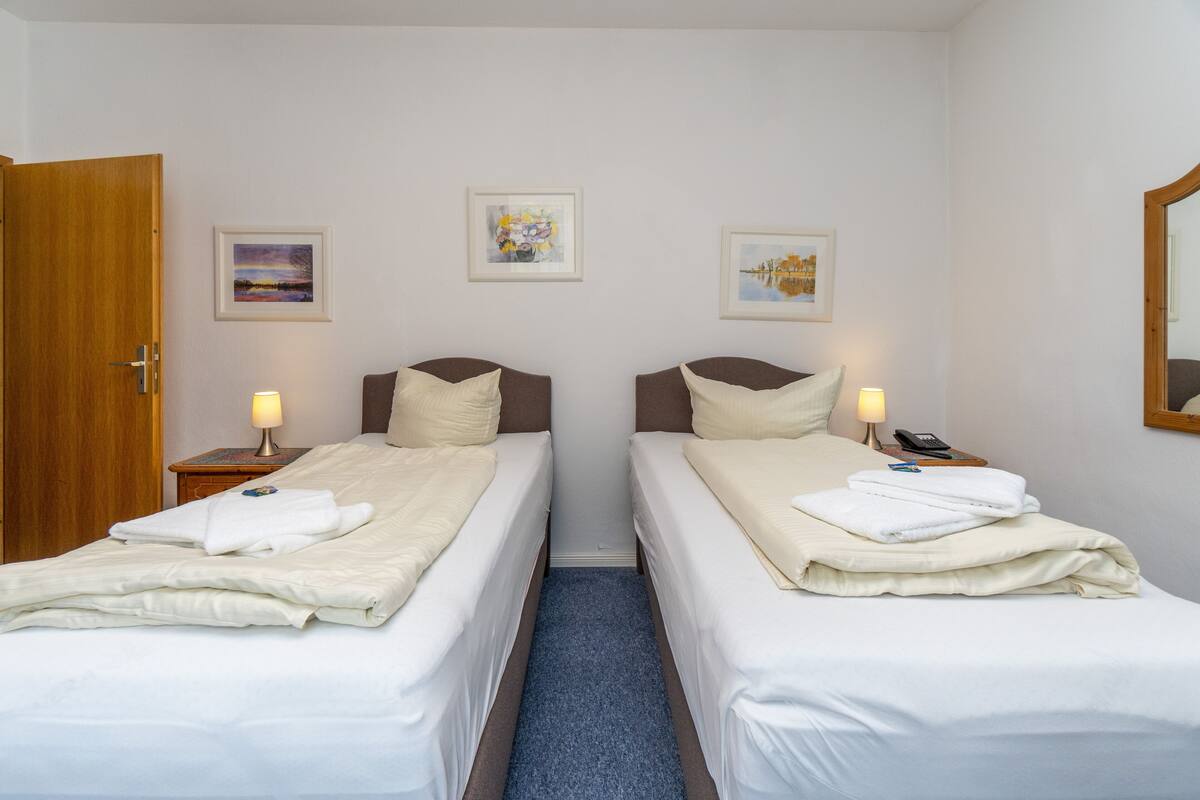 A hotel room with two single beds, a wooden door, a nightstand with a lamp, and framed artwork on the wall.