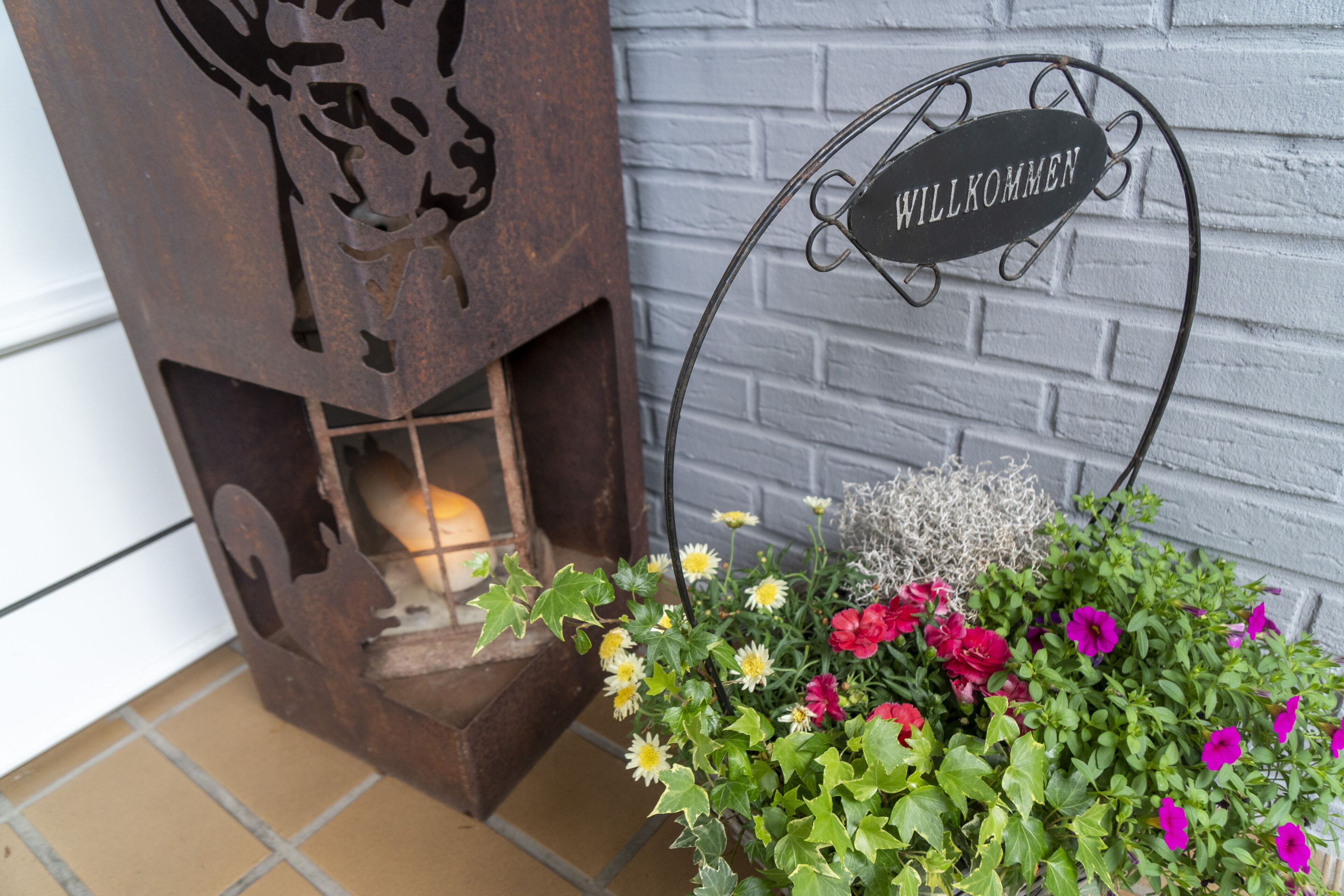 Ein rustikales Metallschild mit "WILLKOMMEN" und einem Blumenarrangement neben einer Backsteinmauer.
