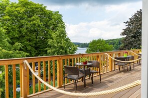 A wooden deck with wicker furniture and rope barriers, overlooking a river and dense greenery. - Hotel Holsteinische Schweiz (Malente)