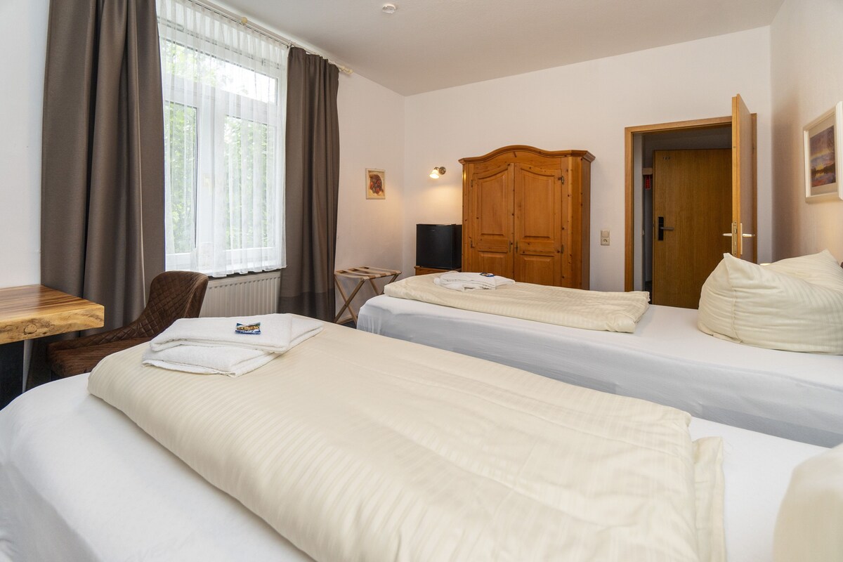 A hotel room with two beds, a wooden wardrobe, a desk with a chair, a television, and a window with curtains.