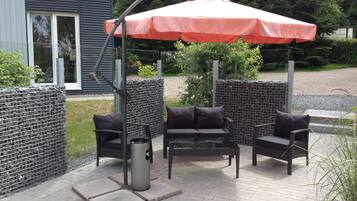 An outdoor patio with a metal grill, two black cushioned chairs, and a red umbrella.
