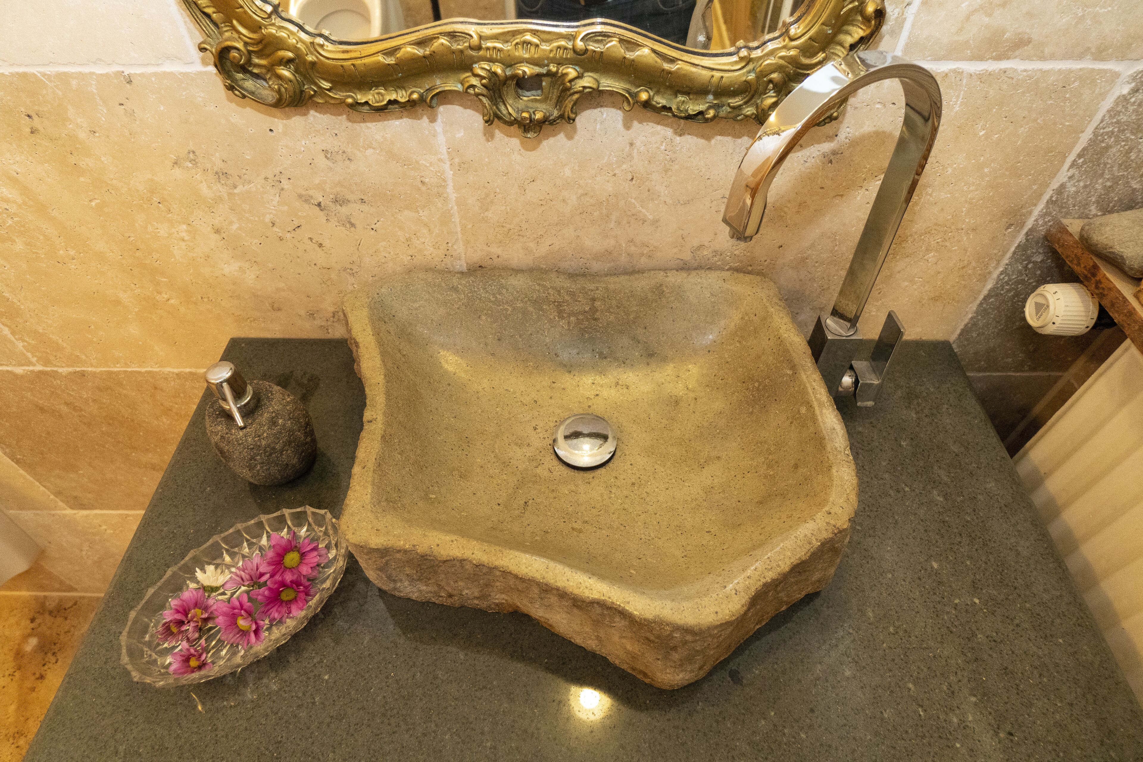 Un baño con un espejo grande y ornamentado, un lavamanos de piedra y un pequeño cuenco decorativo con flores.