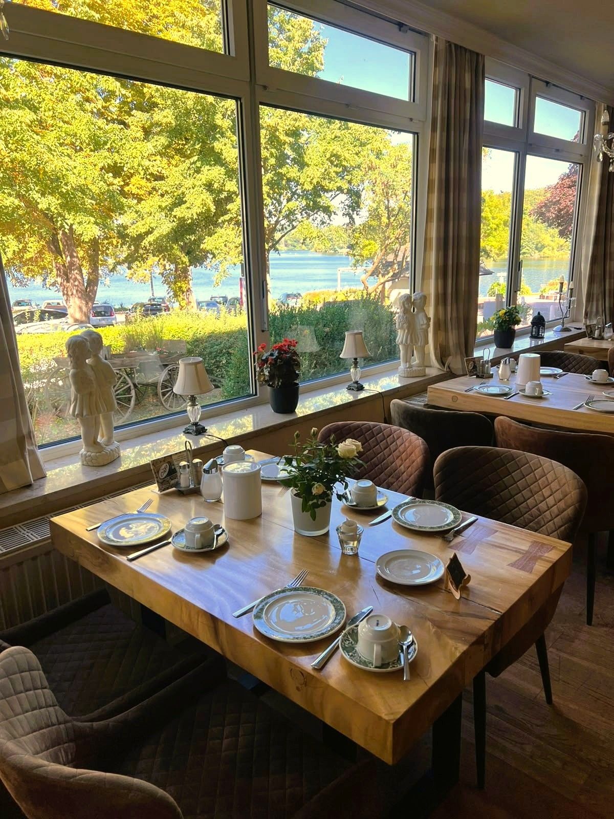 A dining area with tables set for two, large windows with a view of trees and a body of water, and a statue on the windowsill.
