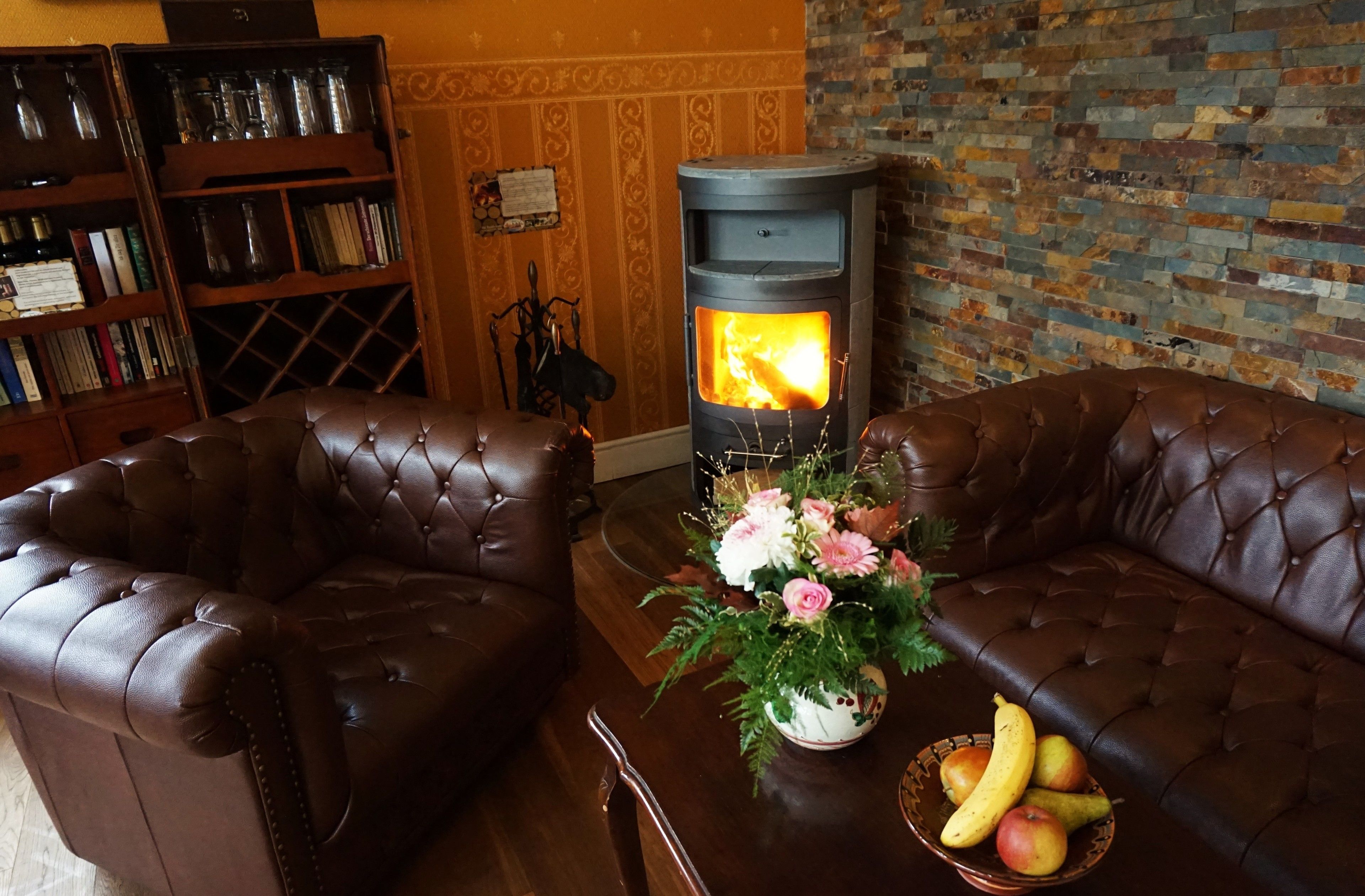 A cozy living room with leather sofas, a wood-burning stove, a bookshelf, and a vase with flowers.