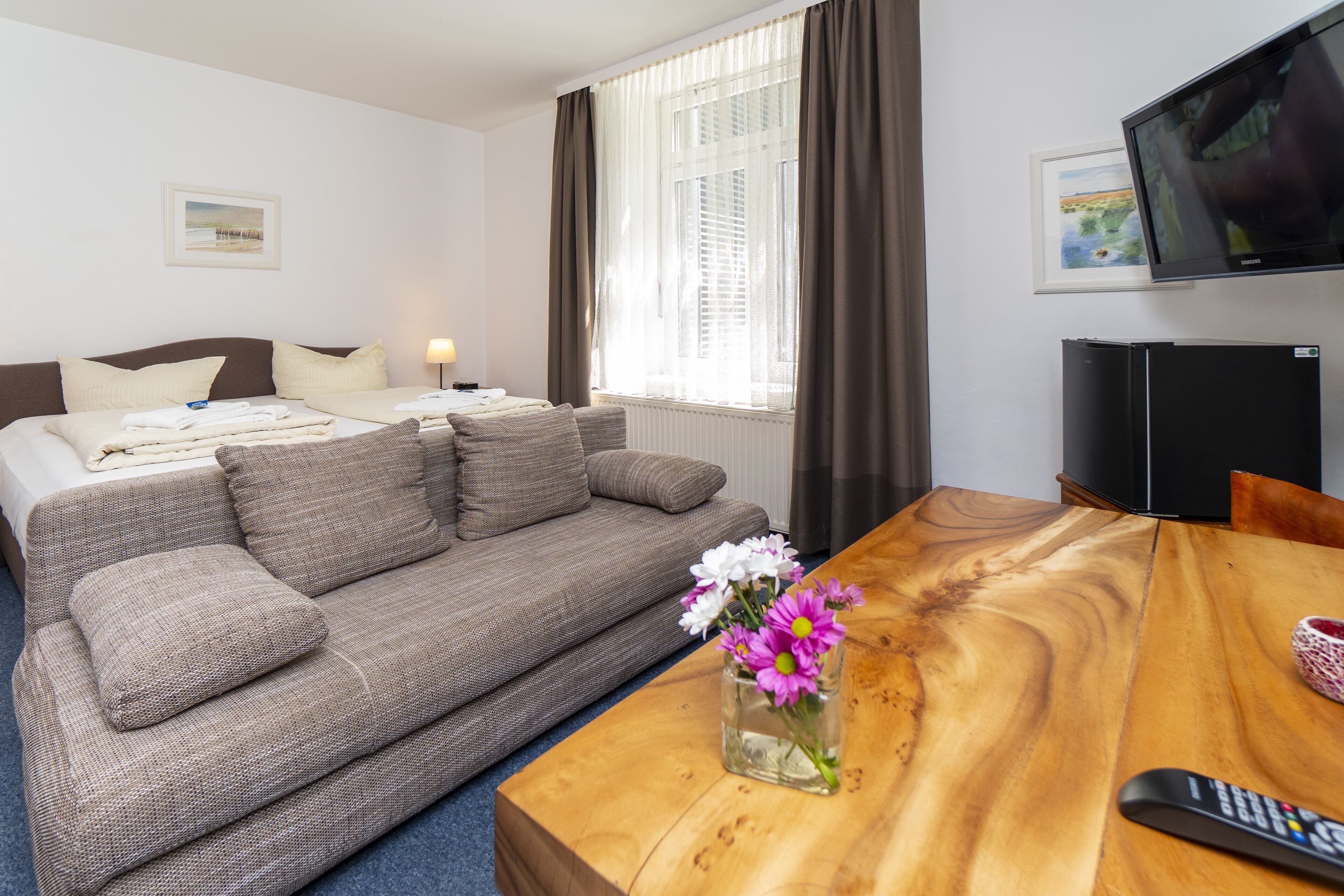 A hotel room with a sofa bed, a wooden coffee table with a vase of flowers, a flat-screen TV, and a framed picture on the wall.