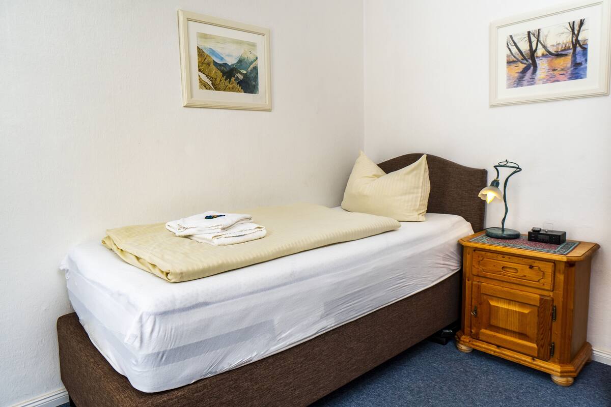 A neatly made bed with a white comforter and a beige blanket, a wooden nightstand with a lamp, and two framed pictures on the wall.