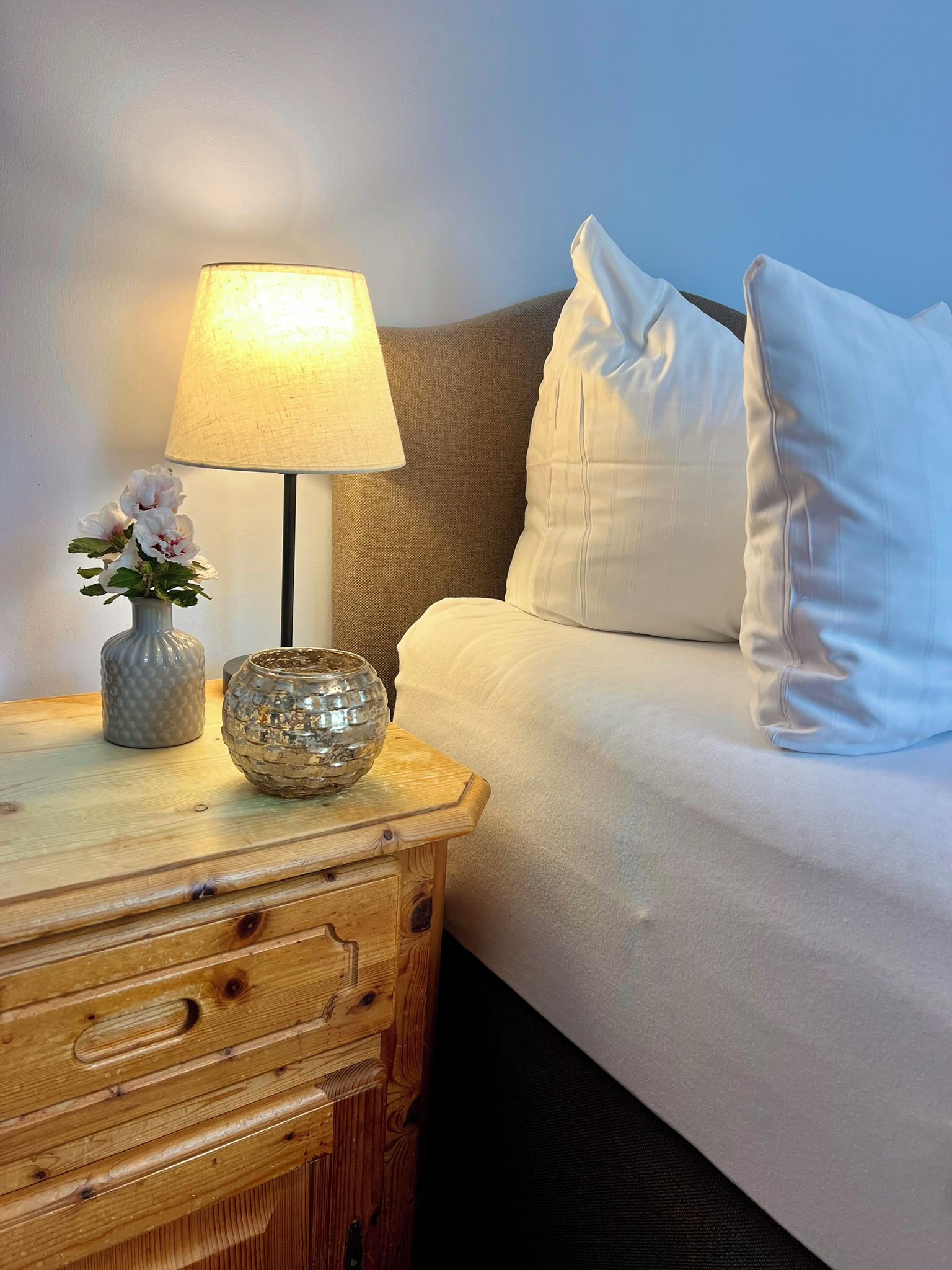 Cama bem arrumada com roupa de cama branca, abajur sobre uma mesa de cabeceira de madeira e um vaso com flores.