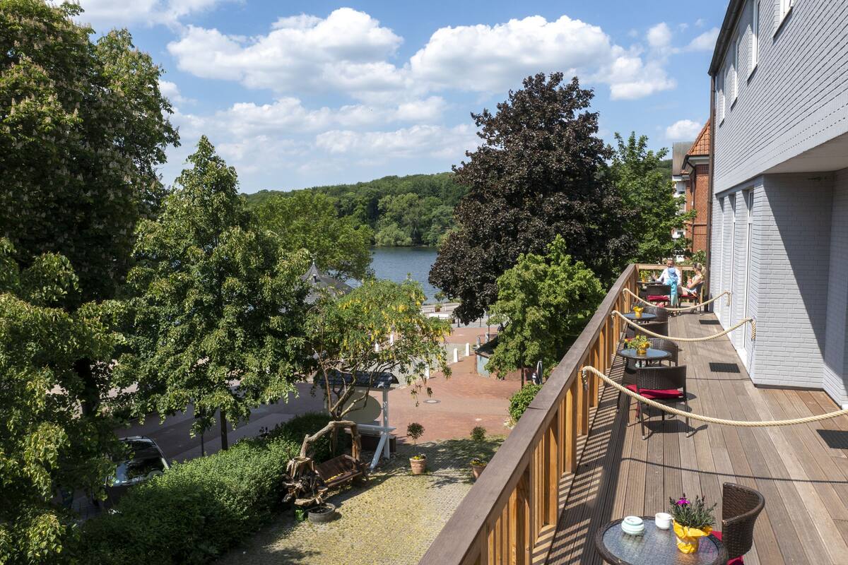 A deck with a view of a lake, surrounded by trees and a building.