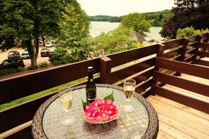 Ein Balkon mit einem Korbstuhl, einer Weinflasche und einem Weinglas mit Blick auf einen Fluss und parkende Autos.