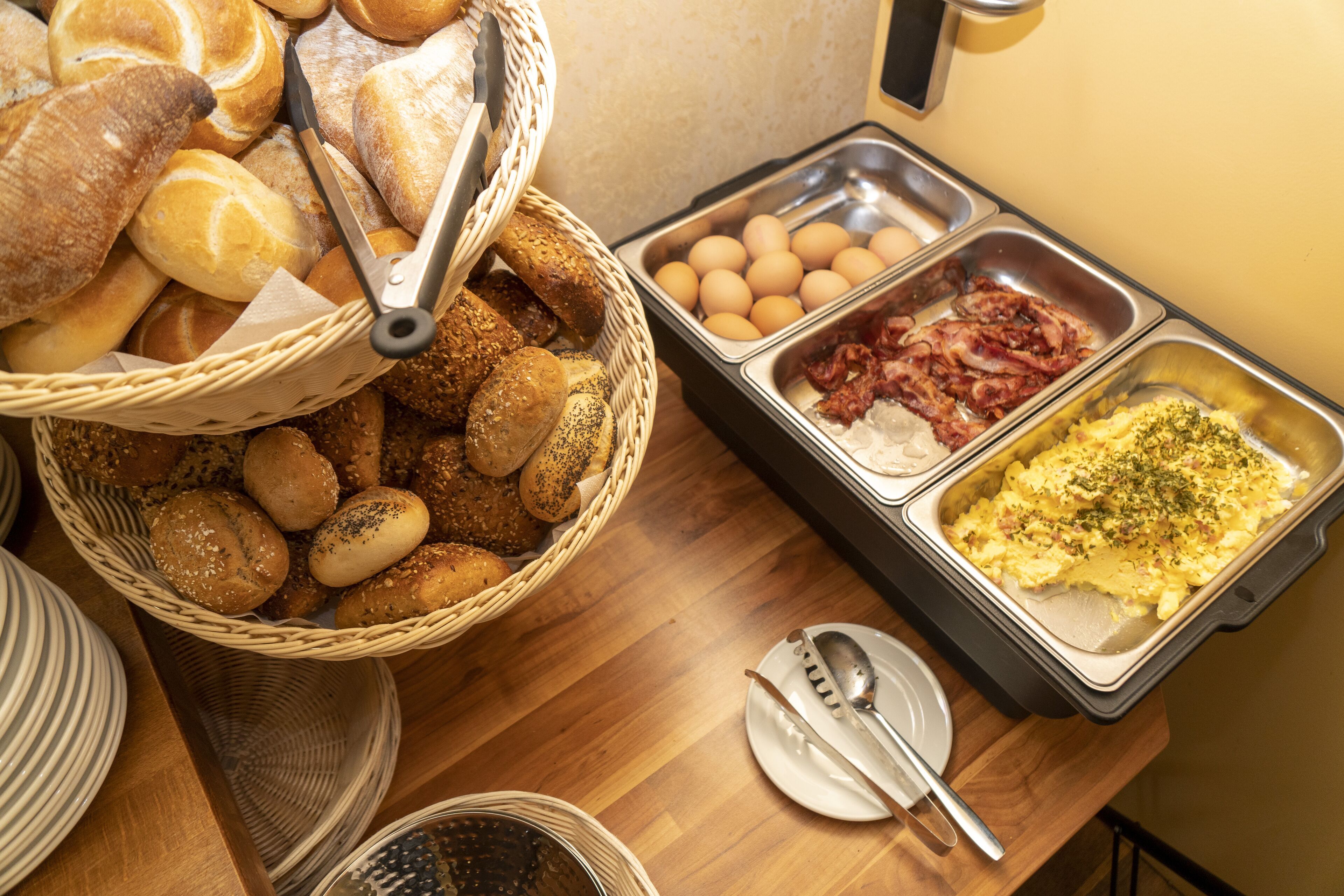 Une table de buffet proposant divers aliments, notamment du pain, des œufs, du bacon et un plat agrémenté d’herbes.