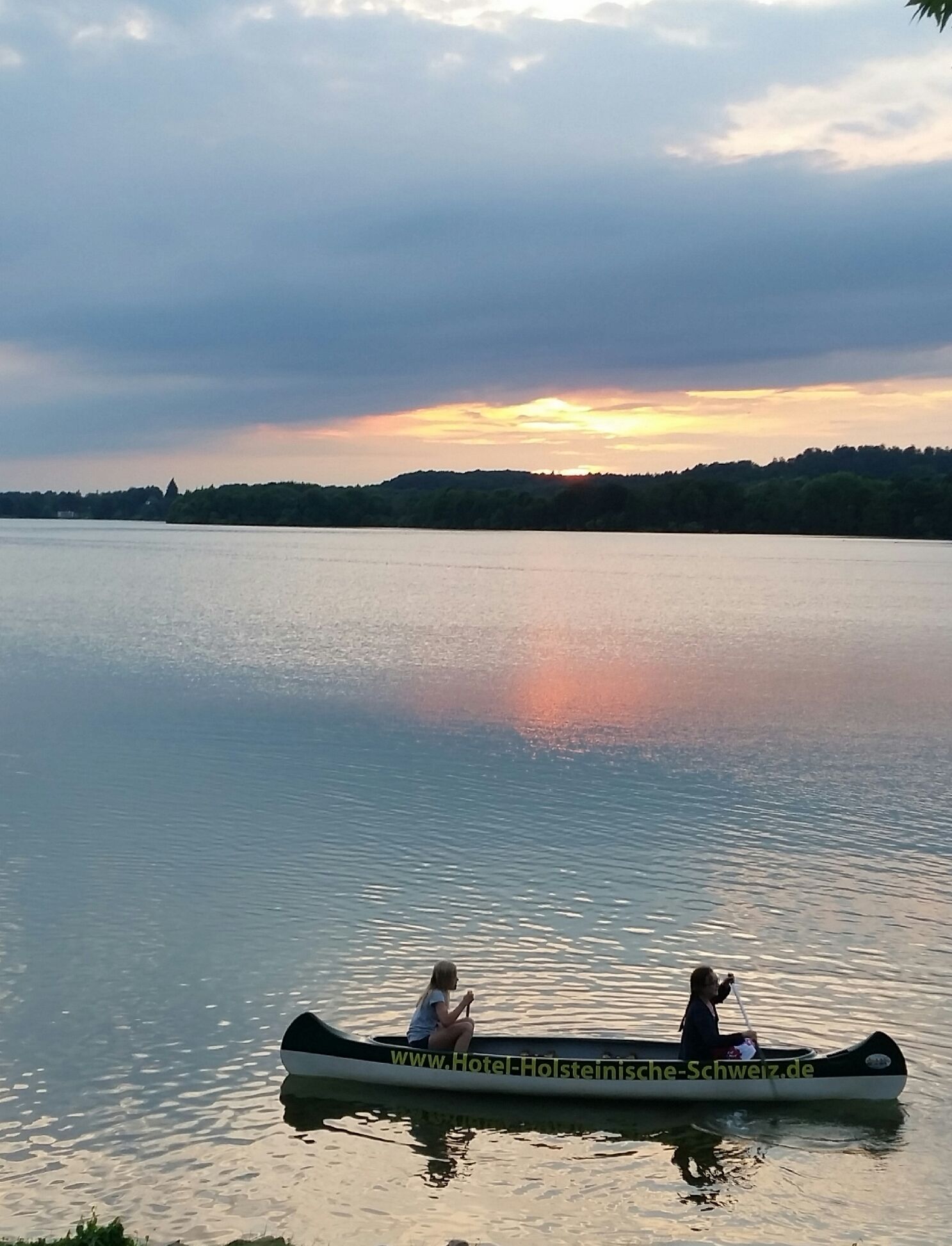 Un lago tranquilo con un atardecer al fondo. Dos personas están en una canoa, una remando.