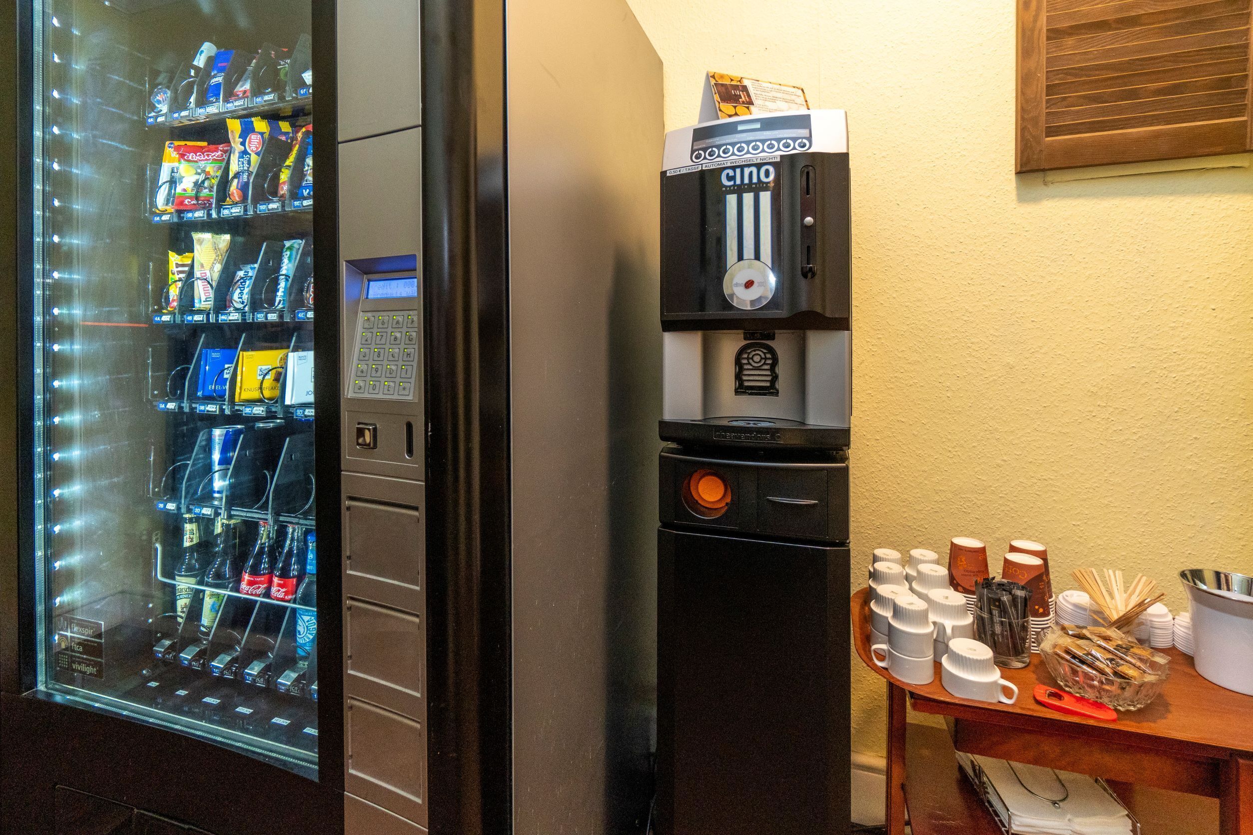 Un distributeur automatique proposant une variété de collations et de boissons, une machine à café et une table avec des tasses et des couverts.