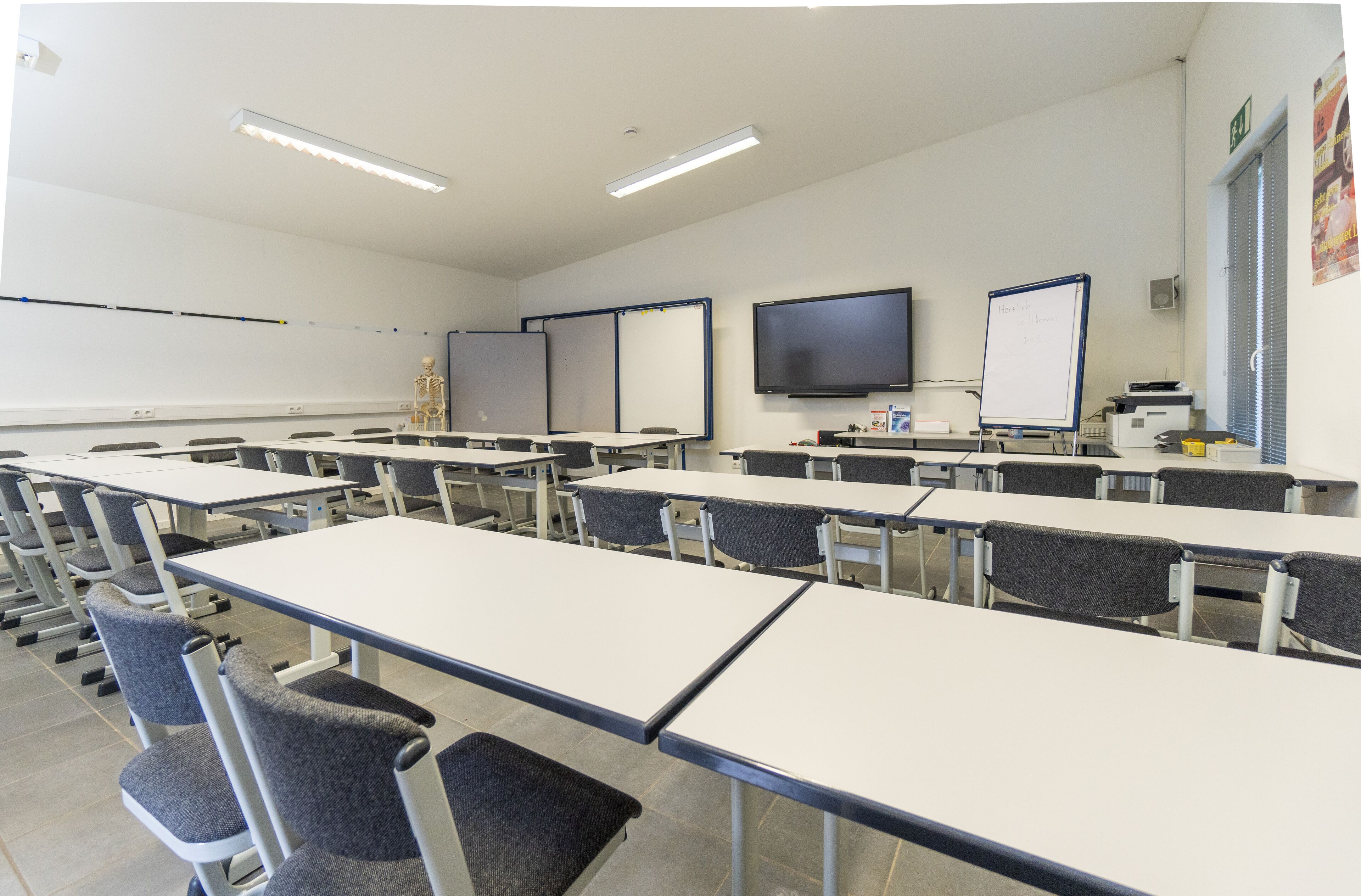 A classroom with rows of desks and chairs, a whiteboard, a projector, and a skeleton model on a table.