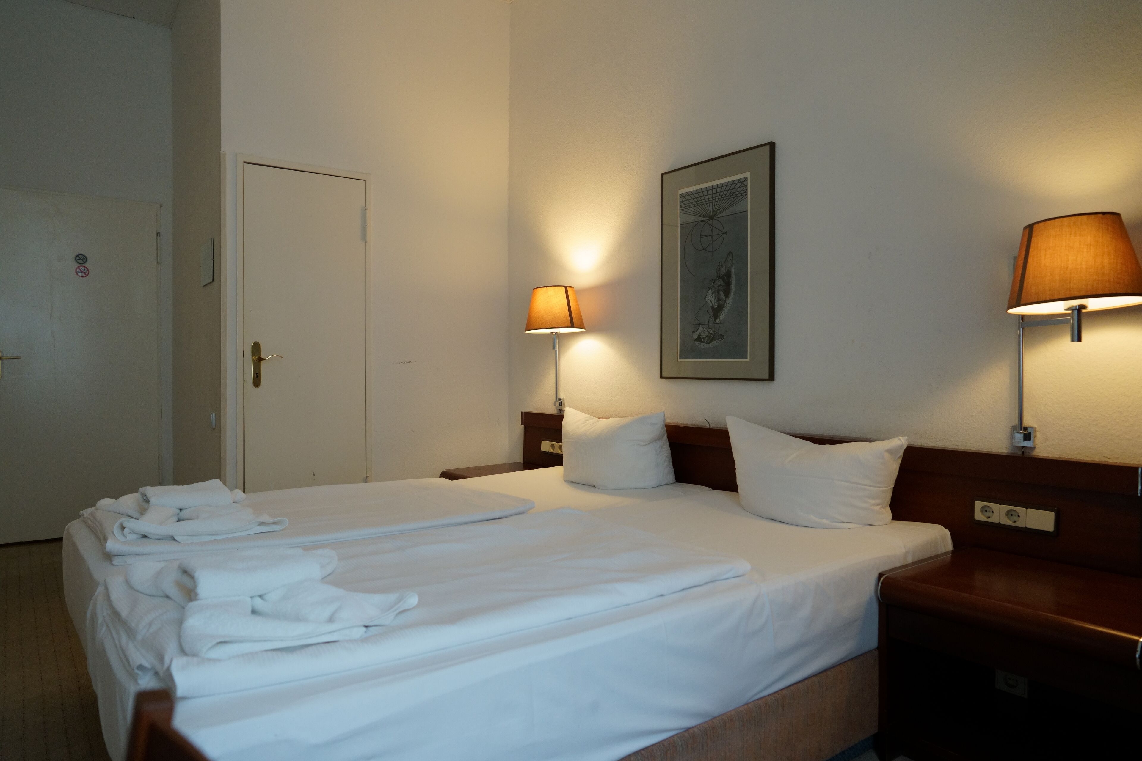 Habitación de hotel con una cama grande, dos mesitas de noche, una lámpara de pared y una puerta.