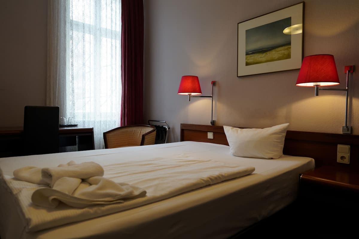 A hotel room with a neatly made bed, two bedside lamps, a chair, a desk, and a window with curtains.