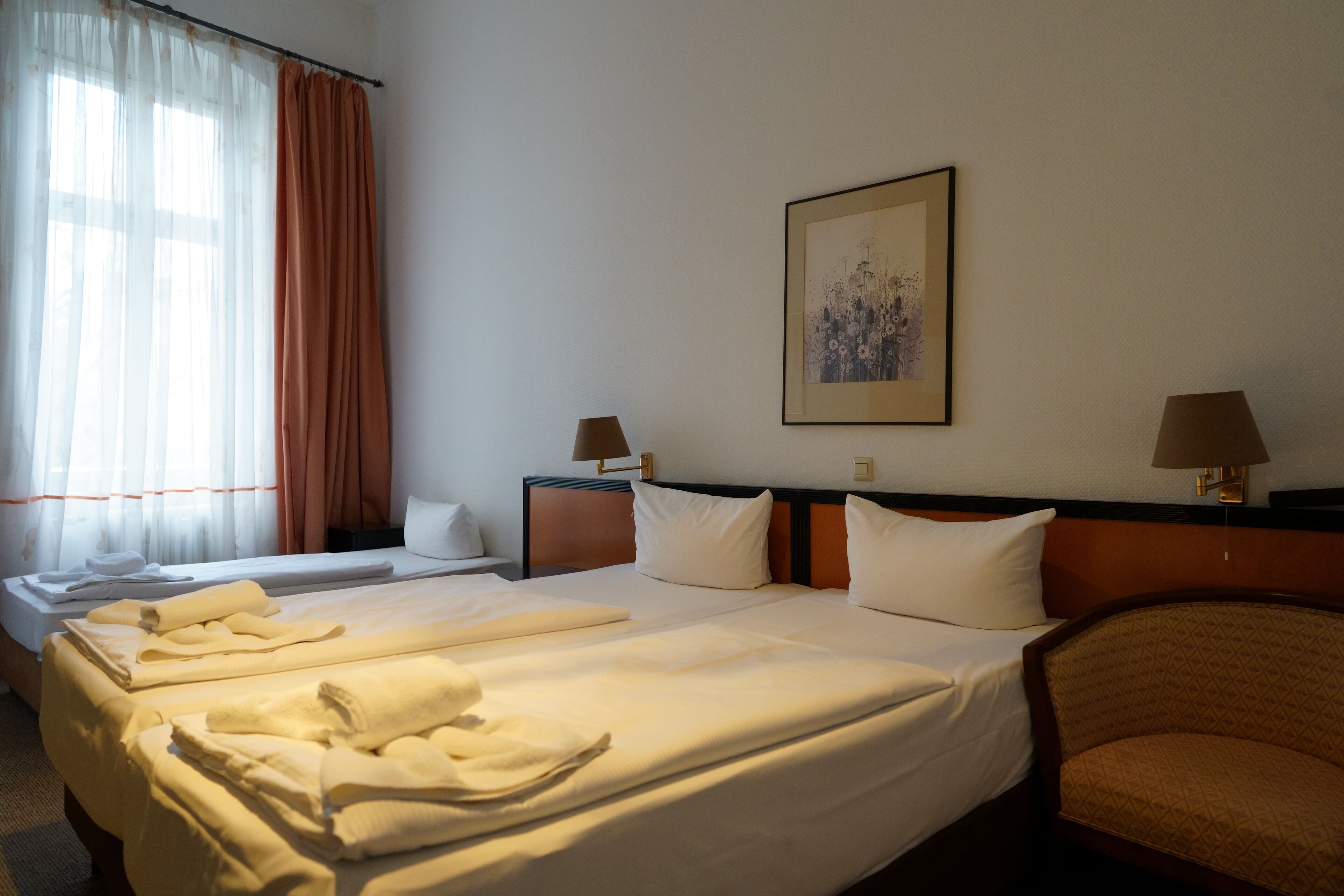 A hotel room with two beds, a large window with sheer curtains, a framed picture on the wall, and two bedside lamps.