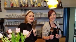 Two people at a bar counter with a pink vase of tulips, stacks of plates labeled "Apfelkuchen," and a variety of bottles and glasses on shelves. - Hotel Cafe Frida (Bredstedt)
