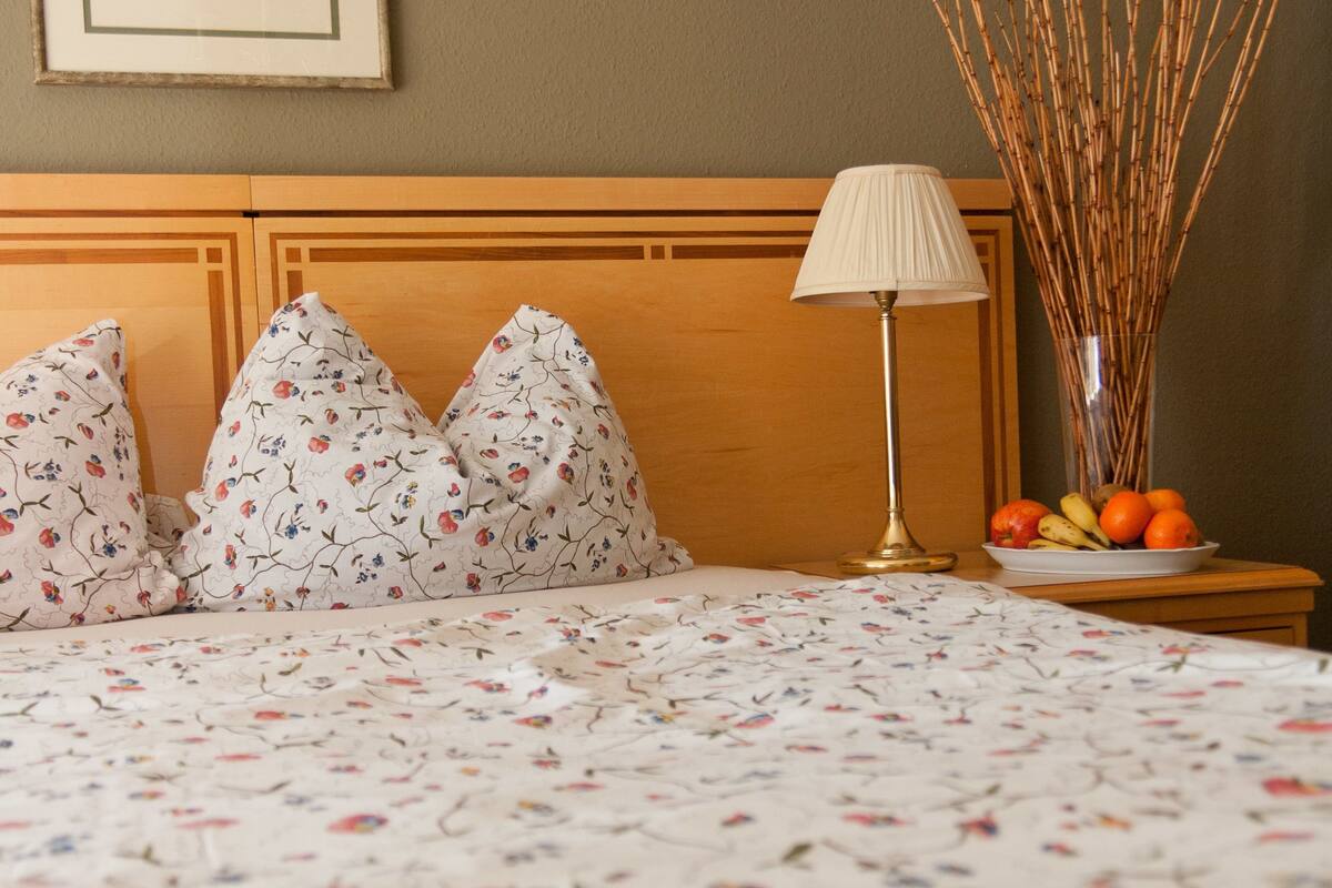 Una cama bien hecha con ropa de cama floral, una lámpara de noche y un florero con ramas y frutas sobre una mesita de noche.