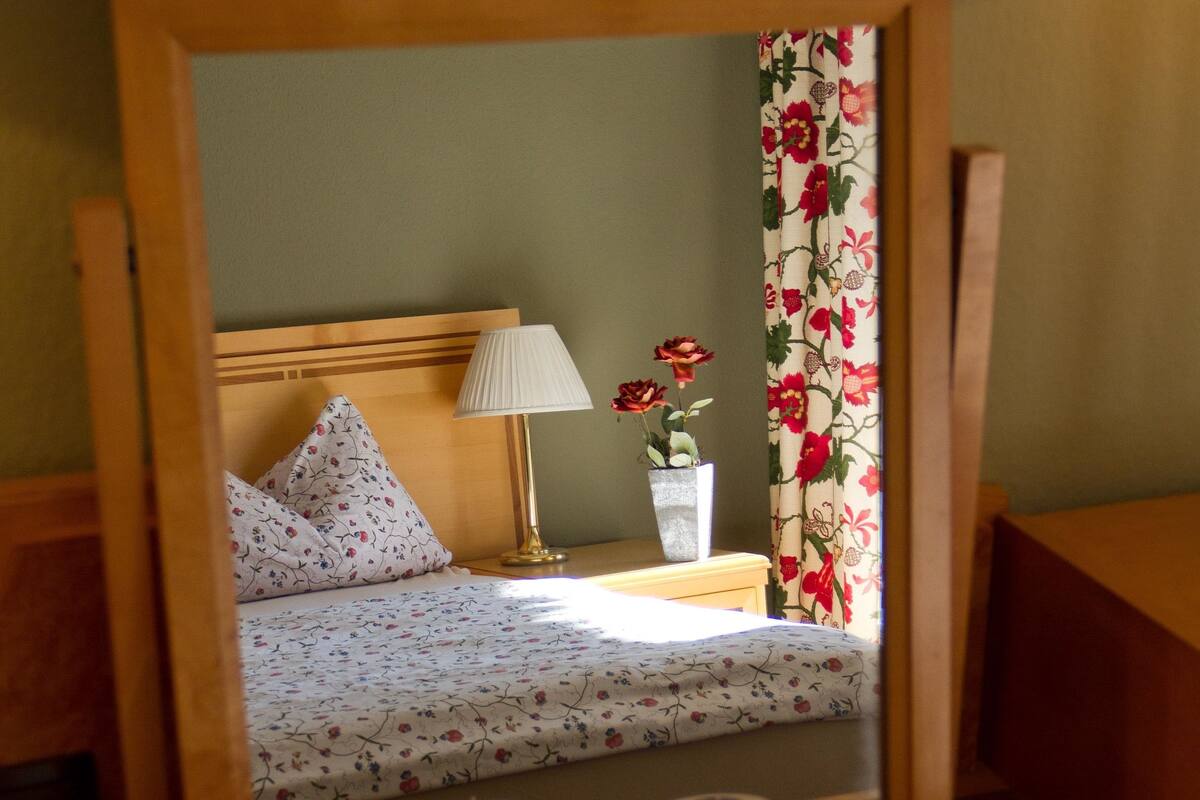 Un dormitorio con cabecera de madera, cama con ropa de cama floral, mesita de noche con lámpara, un florero con flores y un espejo.