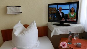 Habitación de hotel con una cama, un televisor sobre un soporte, una botella de agua, un vaso y una caja pequeña sobre una mesa.