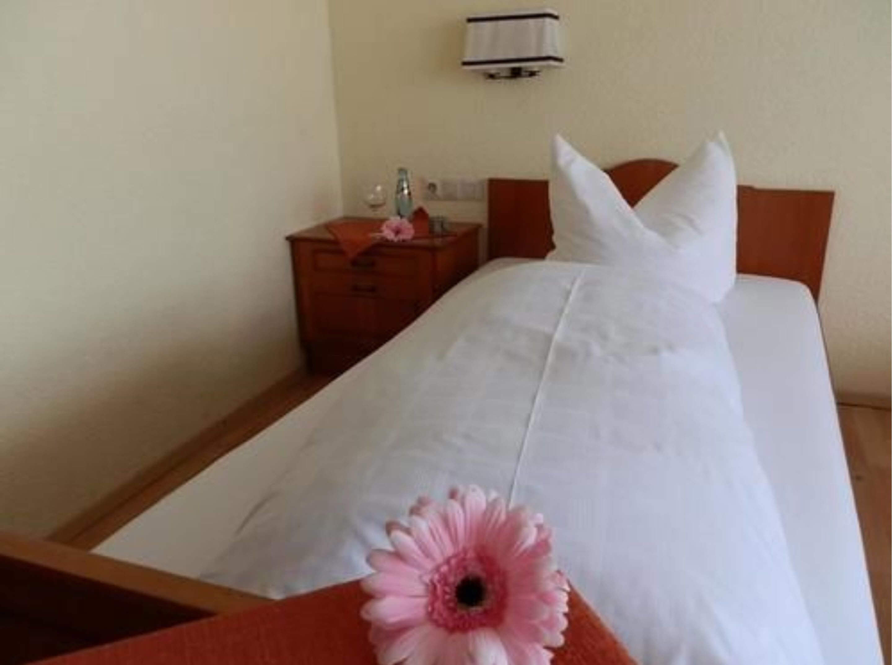 Quarto de hotel com uma cama bem arrumada, uma mesa de cabeceira de madeira com abajur e luminária na parede.