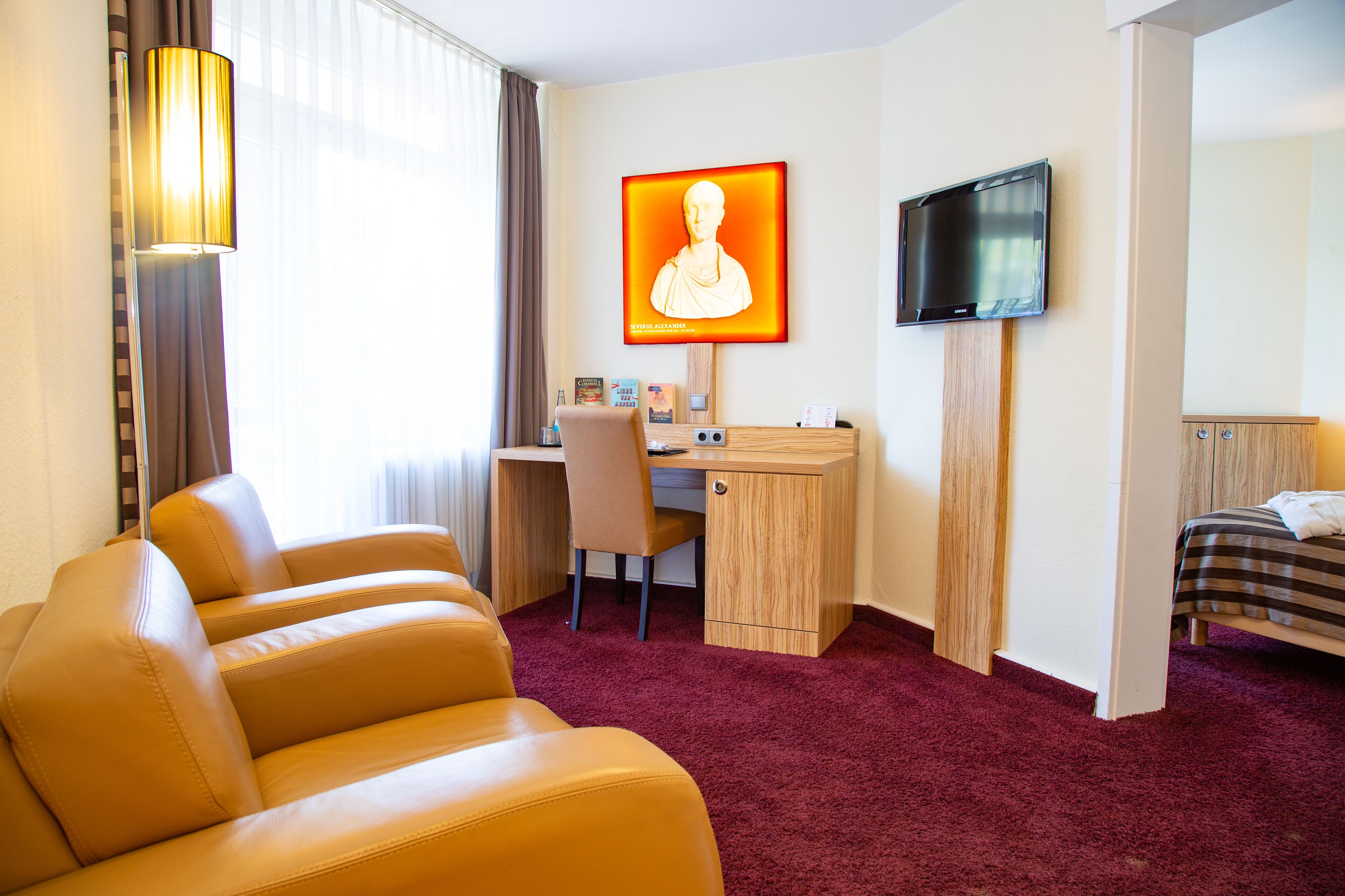 Une chambre d’hôtel équipée d’un bureau, d’une chaise, d’un lit, d’une télévision et d’un grand portrait accroché au mur.
