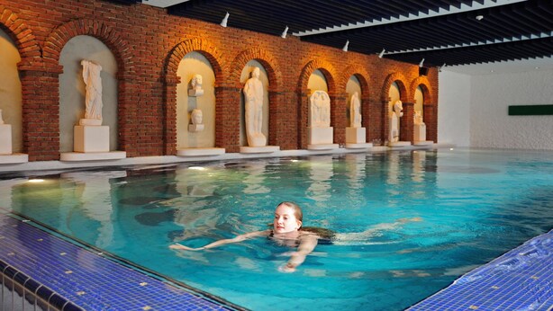 Una piscina con un niño nadando, rodeada de arcos de ladrillo y estatuas.