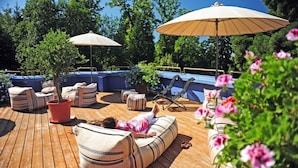 Terraza de madera con mobiliario exterior, incluyendo sillones acolchados y una zona de descanso, rodeada de plantas en macetas y sombrillas.