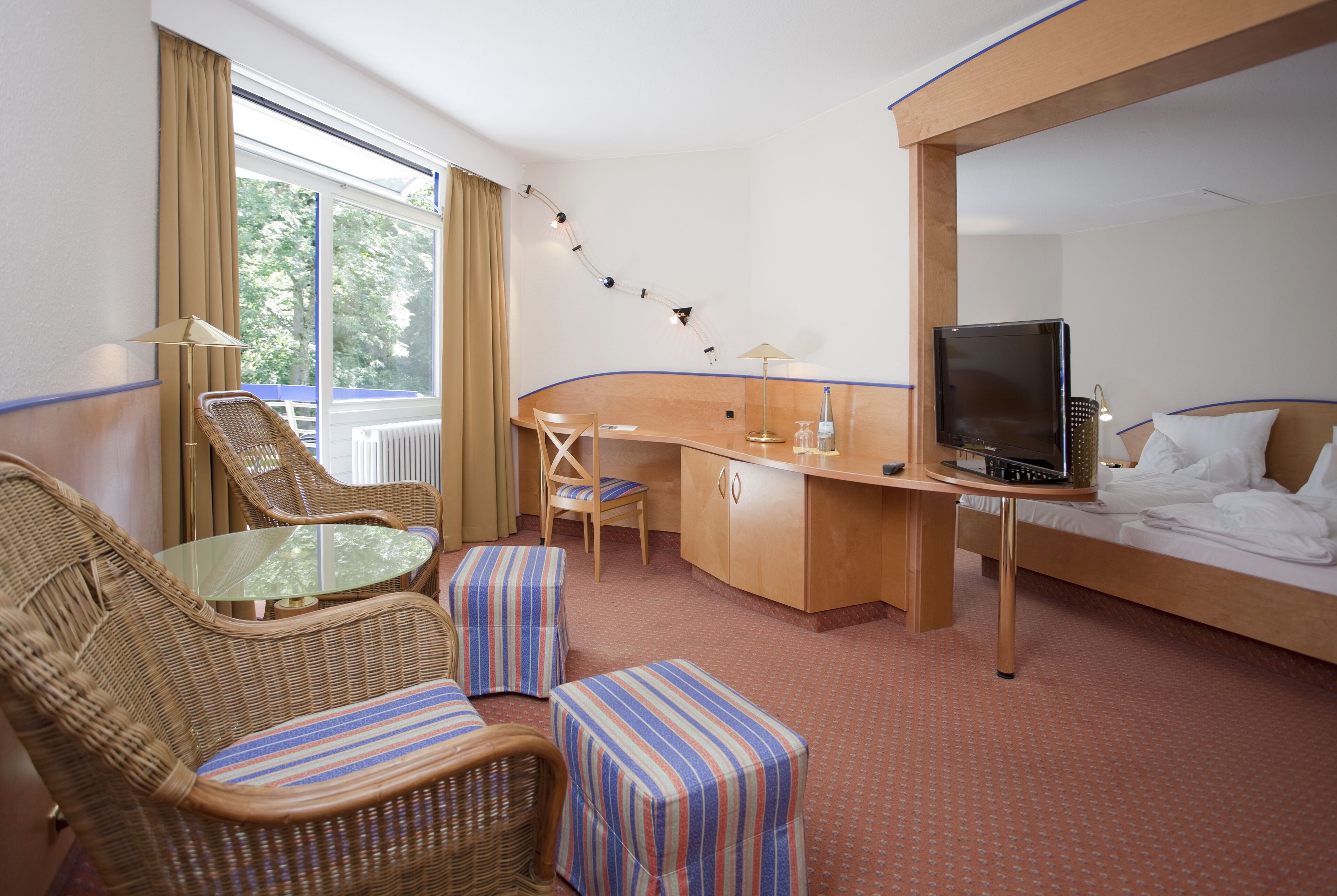A hotel room with a bed, a desk, a television, wicker furniture, and a view of the outdoors.
