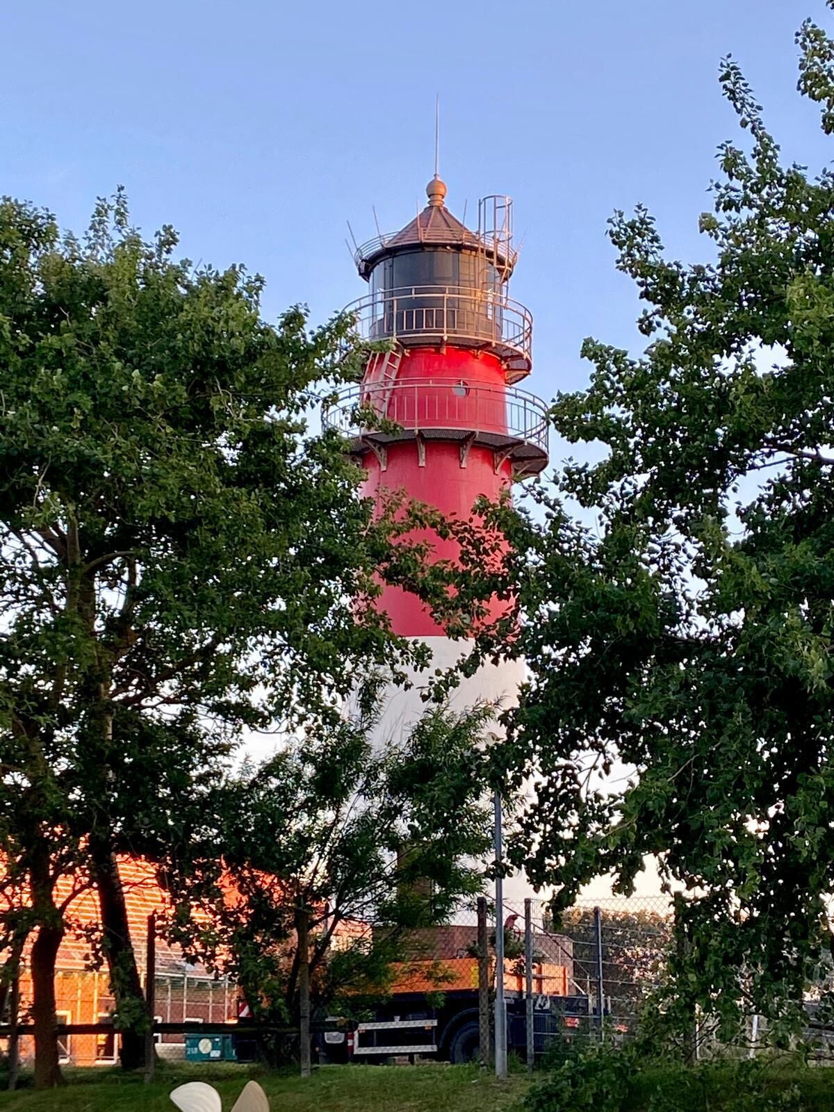A lighthouse with a red and black exterior, surrounded by trees and a fence.