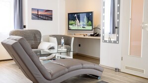 A modern living room with a curved sofa, a glass coffee table, a flat-screen TV, and a painting of a sunset over a pier. - Hotel Boeving (Breckerfeld)