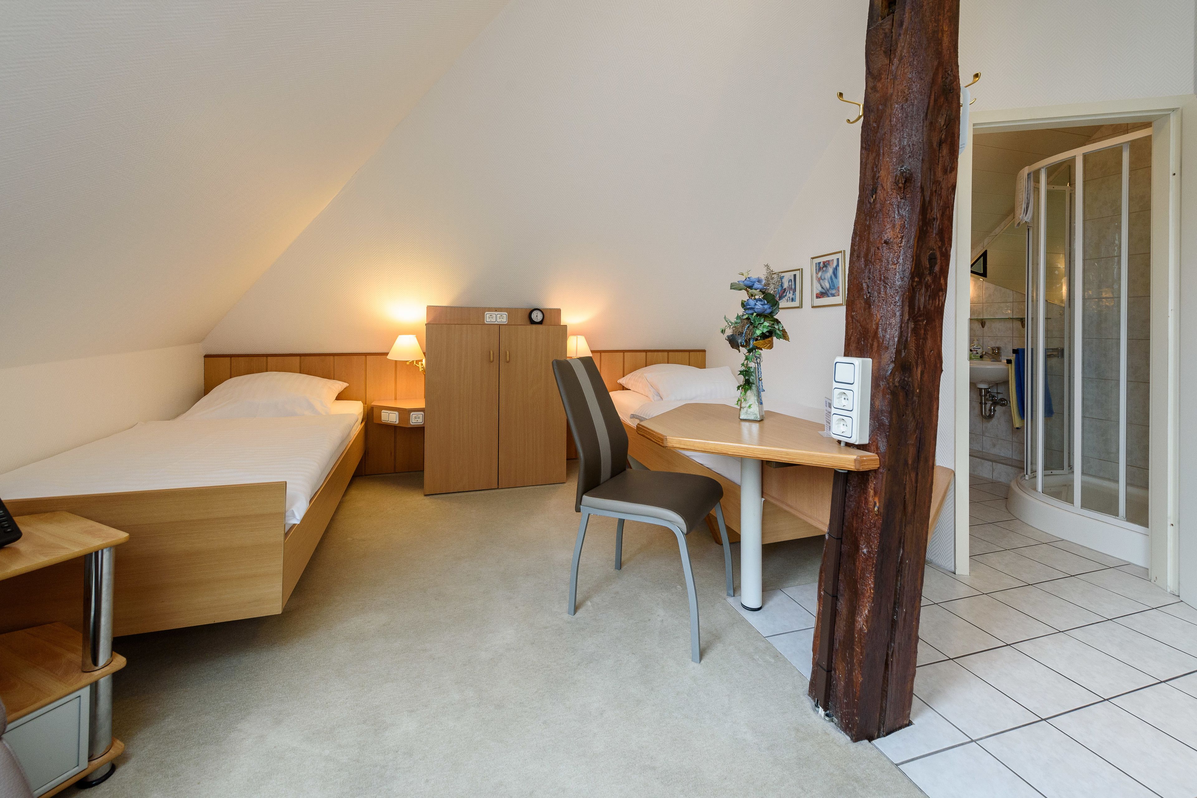 A small, cozy room with two beds, a desk, and a chair. There is a wooden wardrobe and a small table with a vase of flowers.