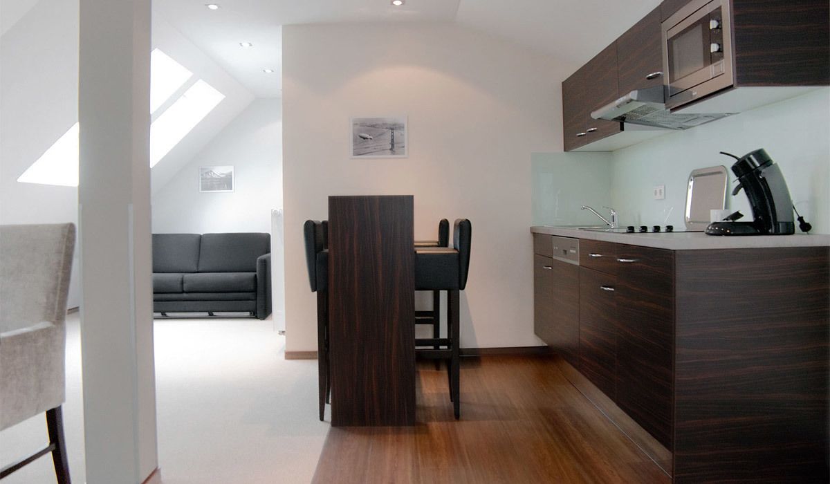 A modern kitchen with dark wood cabinets, a built-in microwave, and a coffee machine. There is a dining area with a black table and chairs, and a sofa in the background.