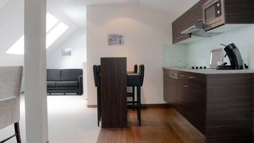 A modern kitchen with dark wood cabinets, a built-in microwave, and a coffee machine. There is a dining area with a black table and chairs, and a sofa in the background.