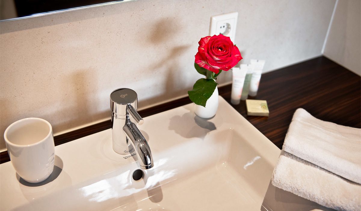 Un lavamanos de baño con un grifo moderno, una taza blanca y un florero con una rosa roja.
