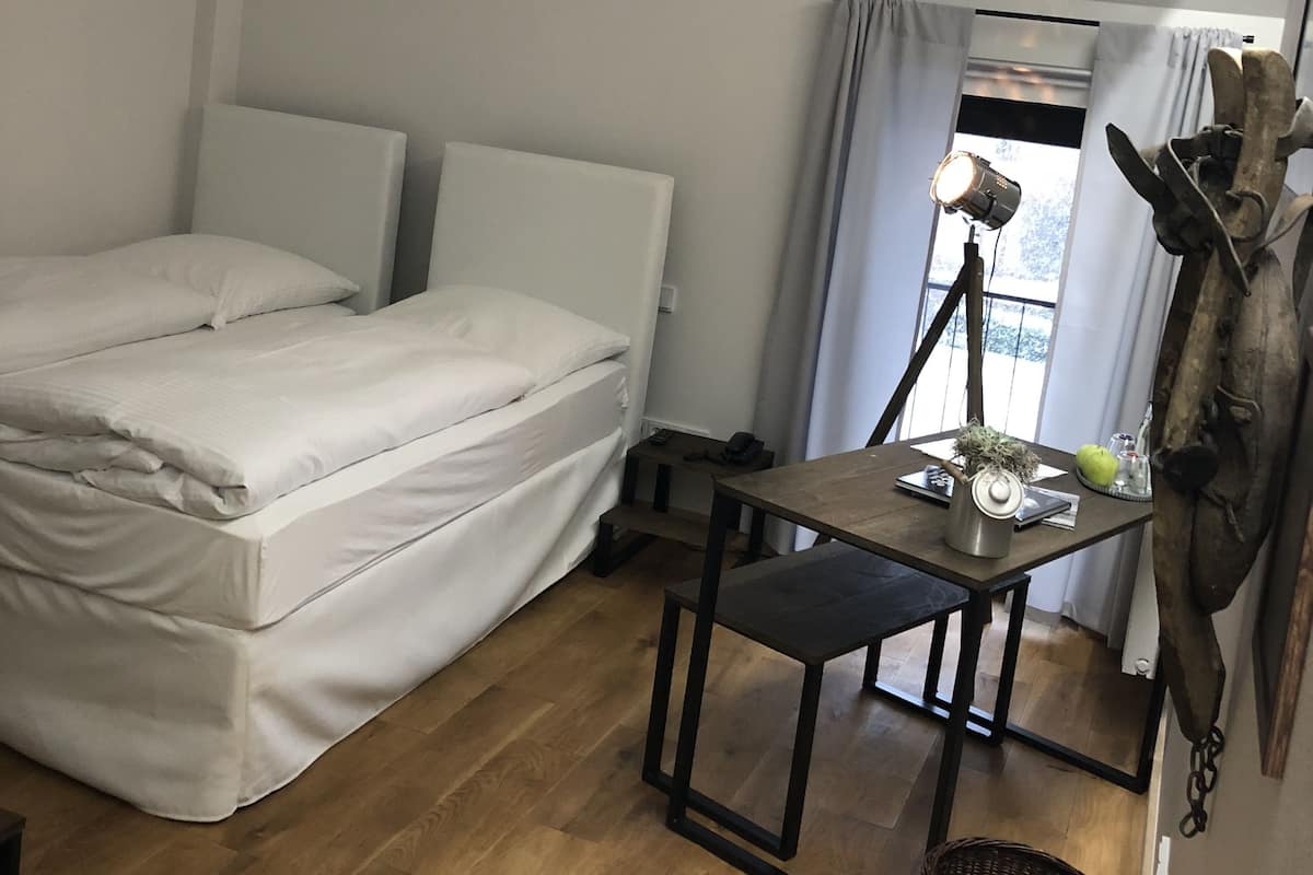 A small, neatly arranged room with a bed, a table with a lamp, and a window with curtains.
