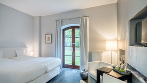 Quarto de hotel com uma cama, uma escrivaninha com luminária, uma cadeira e vista para o exterior através de uma porta.