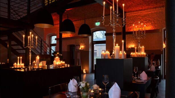 Um restaurante com iluminação suave, mesas iluminadas por velas, um candelabro e uma escadaria ao fundo.