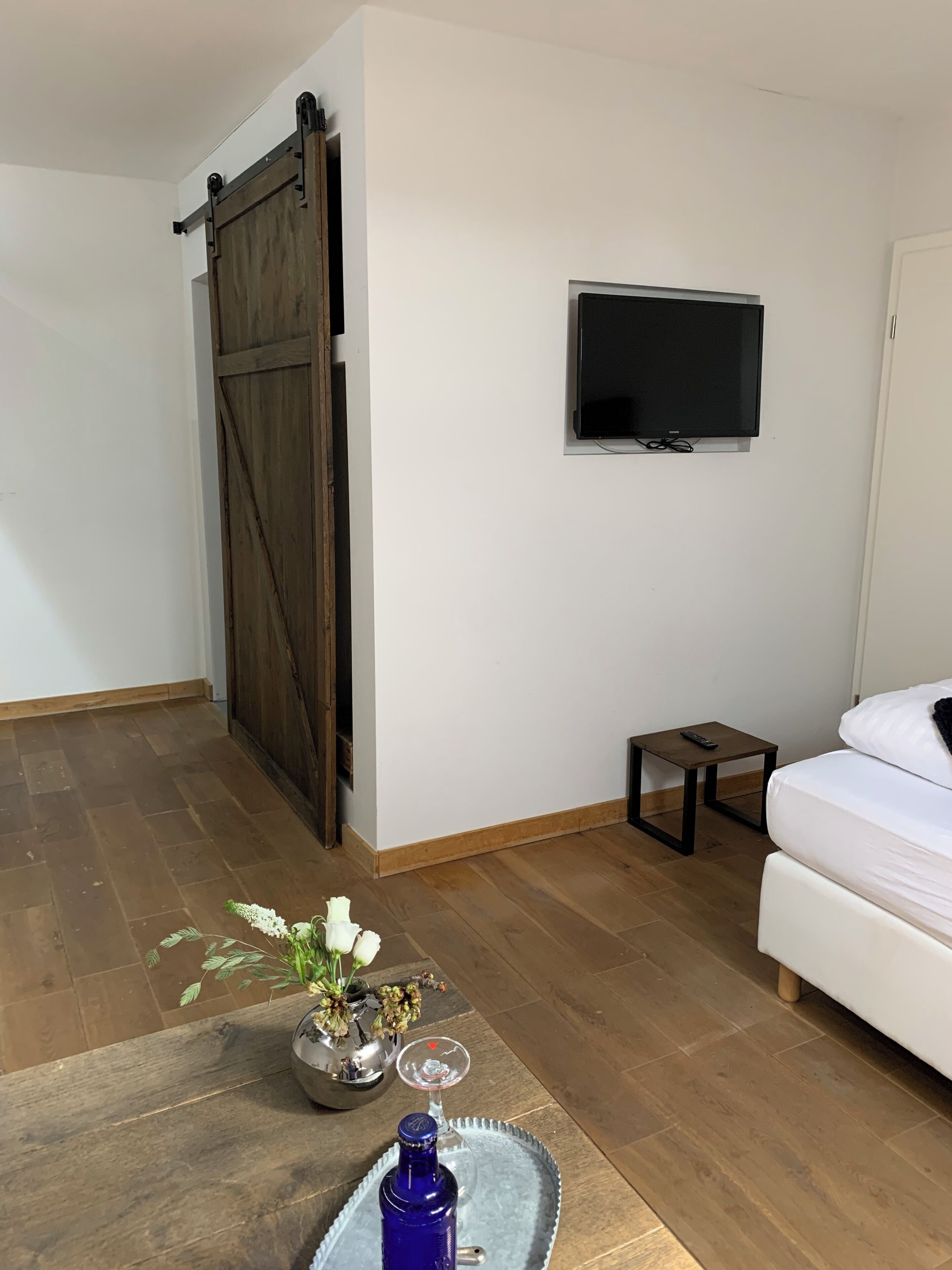 A room with a sliding barn door, a flat-screen TV mounted on the wall, a small bedside table with a remote control, a white bed, and a decorative vase with flowers on a tray.