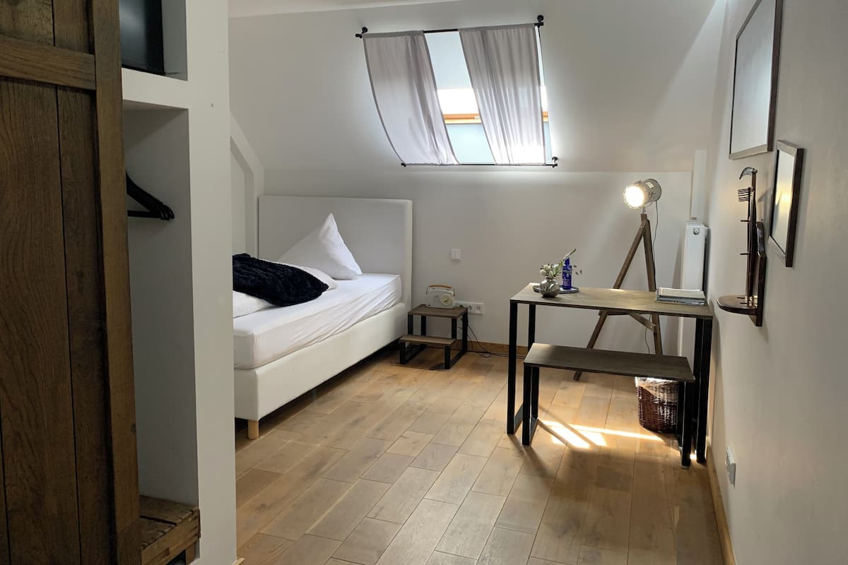 A compact bedroom with a bed, a small table, a desk, a lamp, and a window with curtains.