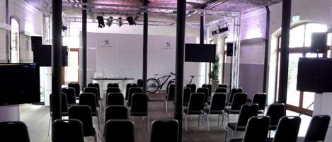 A conference room with rows of chairs, a stage with a bicycle, and large screens.