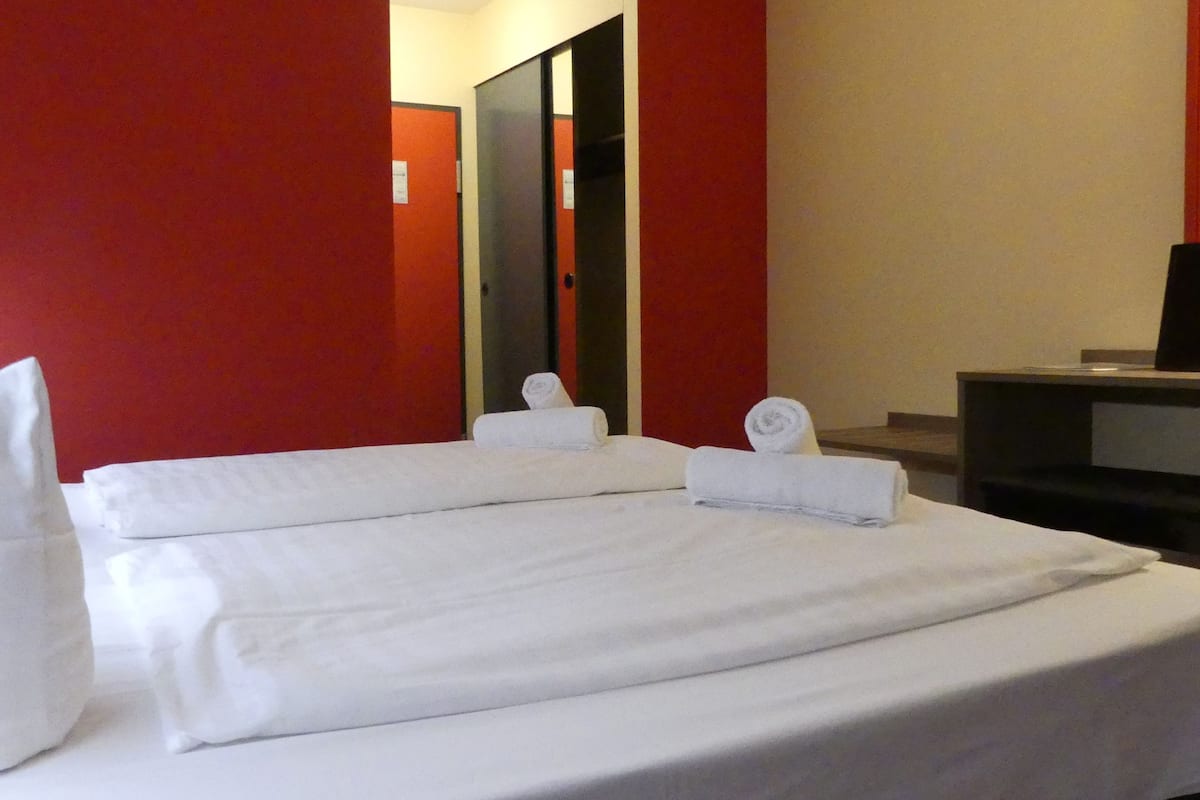 A neatly made bed with white linens and pillows, a television on a stand, and a mirror on the wall.