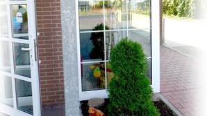 A building entrance with a glass door, a small green bush, and a window reflecting the outside. - Hotel Lindenberger Hof (Ahrensfelde)