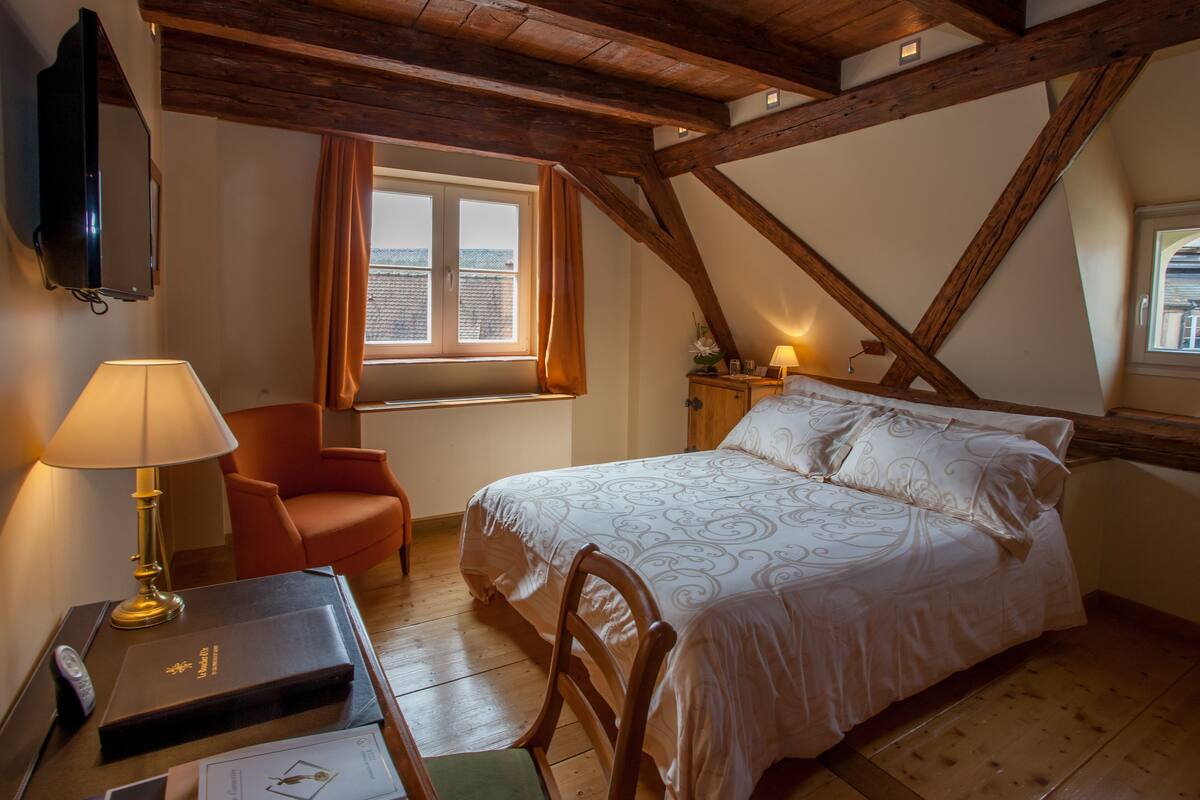 Quarto com cama, cadeira, escrivaninha com luminária, televisão e janela com cortinas.