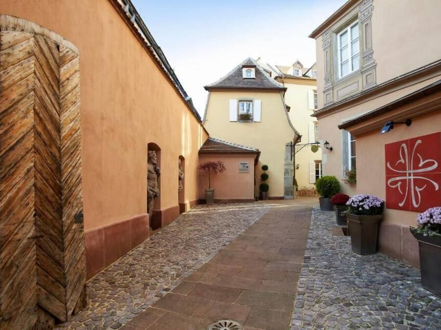 Ein schmaler Kopfsteinpflastergang zwischen Gebäuden, mit einer großen Holztür links und einer roten Wand mit weißem Symbol rechts.